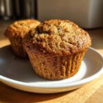 Two golden brown, moist bran muffins sitting on a small white saucer, highlighted by natural sunlight.