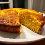 Close-up of a golden brown slice of moist buttermilk cornbread cut from a round loaf on a white plate.