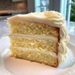 Close-up of a moist, three-layer vanilla cake recipe slice with thick white frosting.
