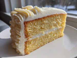 Close-up of a moist slice of vanilla cake recipe layered with white frosting, sitting on a white plate.