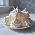 A stack of freshly baked, bright white meringue cookies piped into elegant swirls on a small white plate.