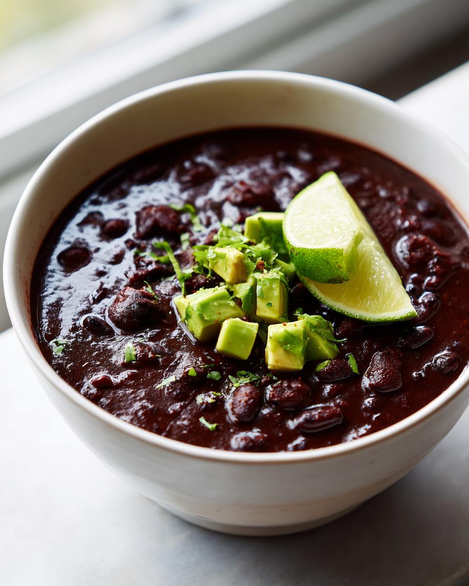 A close-up of a bowl of thick, dark black bean soup garnished with diced avocado and a lime wedge.