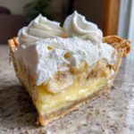 A close-up cross-section of a slice of banana cream pie showing layers of crust, pudding, bananas, and whipped cream.