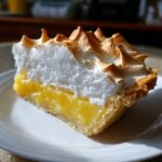 A close-up slice of lemon meringue pie showing bright yellow filling and tall, toasted meringue peaks.