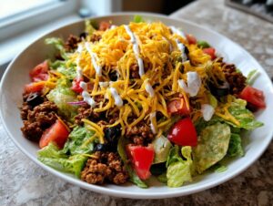 A vibrant bowl of taco salad featuring seasoned ground beef, lettuce, tomatoes, black olives, shredded cheddar, and a drizzle of sour cream.