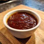Close-up of thick, glossy homemade bbq sauce with visible spice flecks in a small white dipping bowl.
