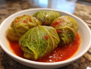 Four plump, green cabbage rolls served in a white bowl with rich tomato sauce.