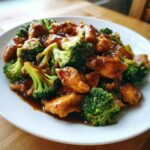 Close-up of glossy chicken and broccoli stir-fry coated in rich brown sauce on a white plate.