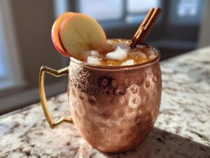 A frosty copper mug filled with an apple cider moscow mule, garnished with apple slices and a cinnamon stick.