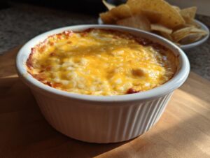 A close-up of hot, baked chili cheese dip topped with melted yellow and white cheese, served in a white ramekin next to tortilla chips.