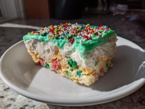 A single slice of Christmas tree cheesecake featuring a colorful cereal base, white filling, green frosting, and rainbow sprinkles.