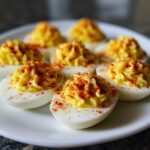 Close-up of creamy deviled eggs piped high and sprinkled with bright red paprika on a white plate.