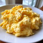 Close-up of fluffy, creamy scrambled eggs piled high on a white plate, ready to eat.