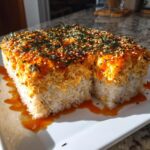 A square portion of baked sushi bake with a creamy orange topping, sprinkled with sesame seeds and seaweed, sitting on a white plate.