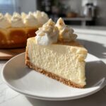 A close-up slice of creamy eggnog cheesecake recipe on a white plate, topped with whipped cream and nutmeg.