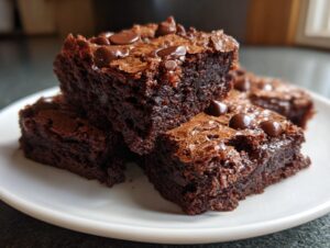 Close-up of several fudgy brownies stacked on a white plate, topped with melted chocolate chips.