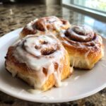 Close-up of three freshly baked cinnamon buns covered in thick white icing, sitting on a white plate.
