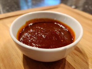 Close-up of a small white bowl filled with thick, dark red homemade bbq sauce sitting on a wooden cutting board.