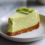 A close-up of a thick slice of bright green key lime cheesecake with a graham cracker crust, topped with lime zest.