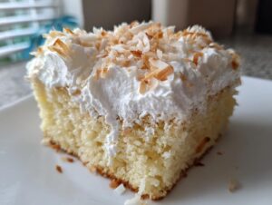 Close-up of a moist slice of coconut poke cake topped with white whipped cream and toasted coconut flakes.