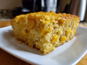 Close-up of a moist, golden slice of cornbread casserole studded with whole corn kernels, served on a white plate.