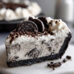 A decadent slice of no-bake ice cream pie featuring a dark cookie crust, creamy filling mixed with cookie pieces, and topped with whipped cream and whole cookies.
