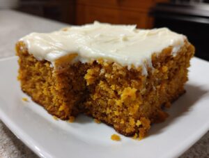 A close-up of a thick, moist square of pumpkin bars with cream cheese frosting on a white plate.