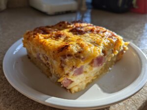 A thick, square slice of cheesy scalloped potatoes and ham casserole with a golden-brown, baked top, served on a white plate.