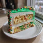 A tall slice of Christmas tree cheesecake featuring layers of funfetti cake, white frosting, and green icing with festive sprinkles.