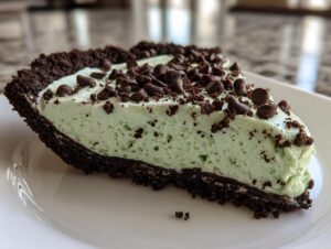 A close-up of a slice of creamy, light green grasshopper pie with a dark chocolate crust, topped with mini chocolate chips.