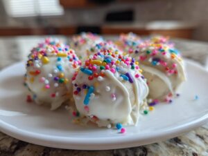 A close-up of several white chocolate coated sugar cookie truffles generously topped with colorful rainbow sprinkles.