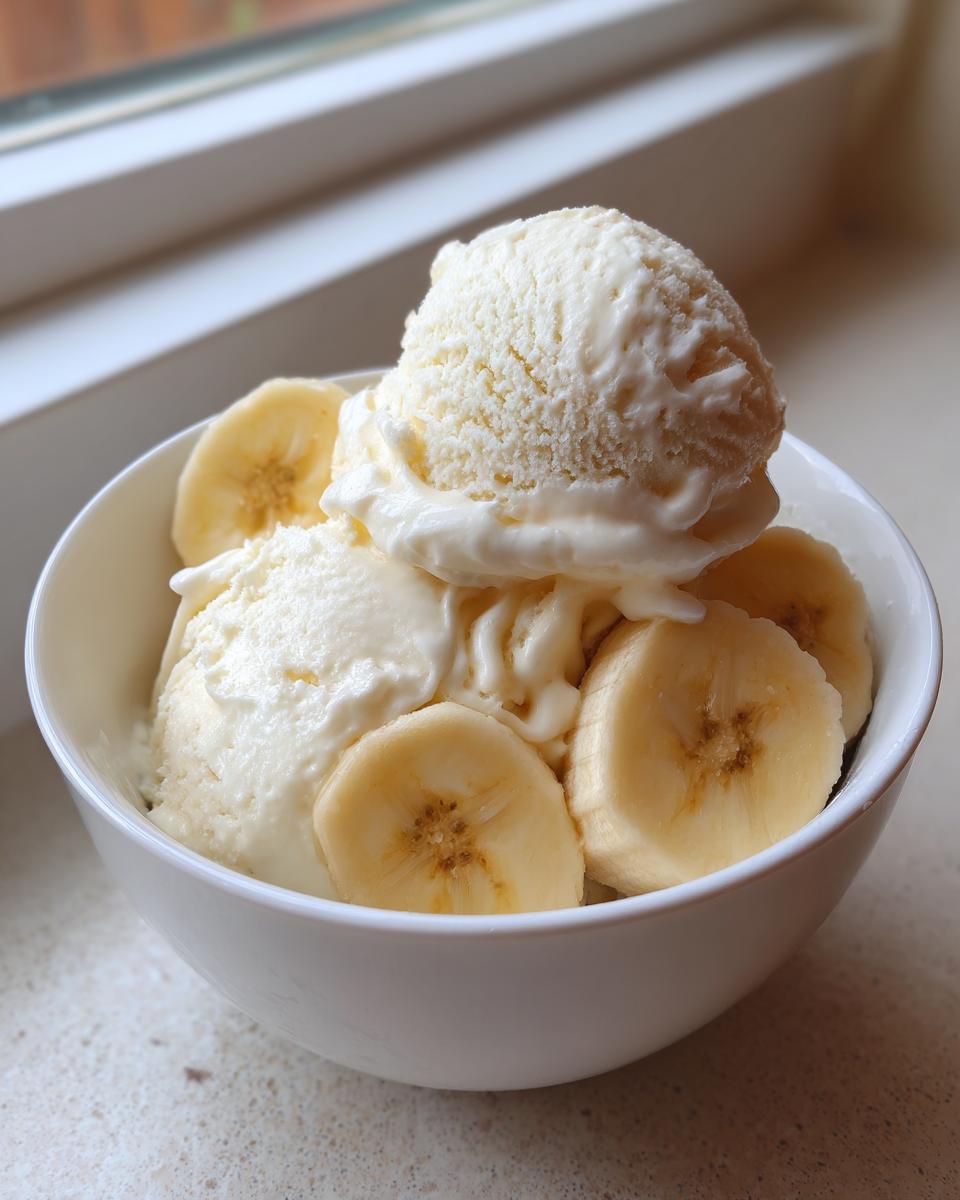 A white bowl filled with scoops of creamy banana ice cream garnished with fresh banana slices.