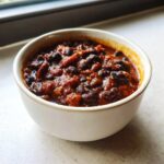 Close-up of a white bowl filled with hearty, rich black bean chili, set near a window.