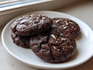 Four rich, dark chocolate Andes mint cookies stacked slightly on a white plate near a window.