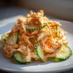 A close-up of a vibrant kani salad featuring shredded imitation crab mixed with a creamy orange dressing, sliced cucumbers, and sprinkled with sesame seeds.