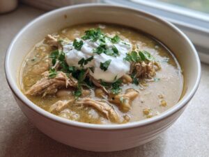 A close-up of a bowl of crockpot white chicken chili, topped with shredded chicken, sour cream, and fresh parsley.