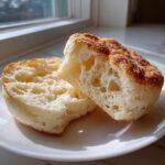 A homemade english muffin split in half, showing its signature nooks and crannies texture on a white plate.