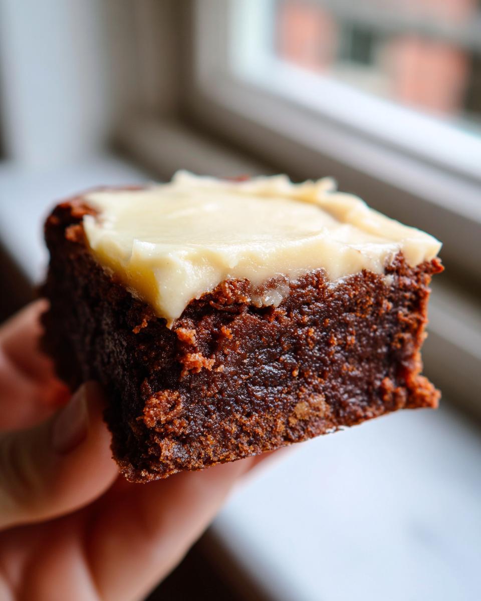 A hand holding a single, moist square of dark gingerbread brownies topped with thick, pale frosting.