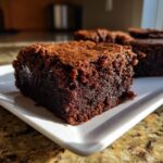 A close-up, sunlit shot of a rich, fudgy square of date brownies resting on a white plate.
