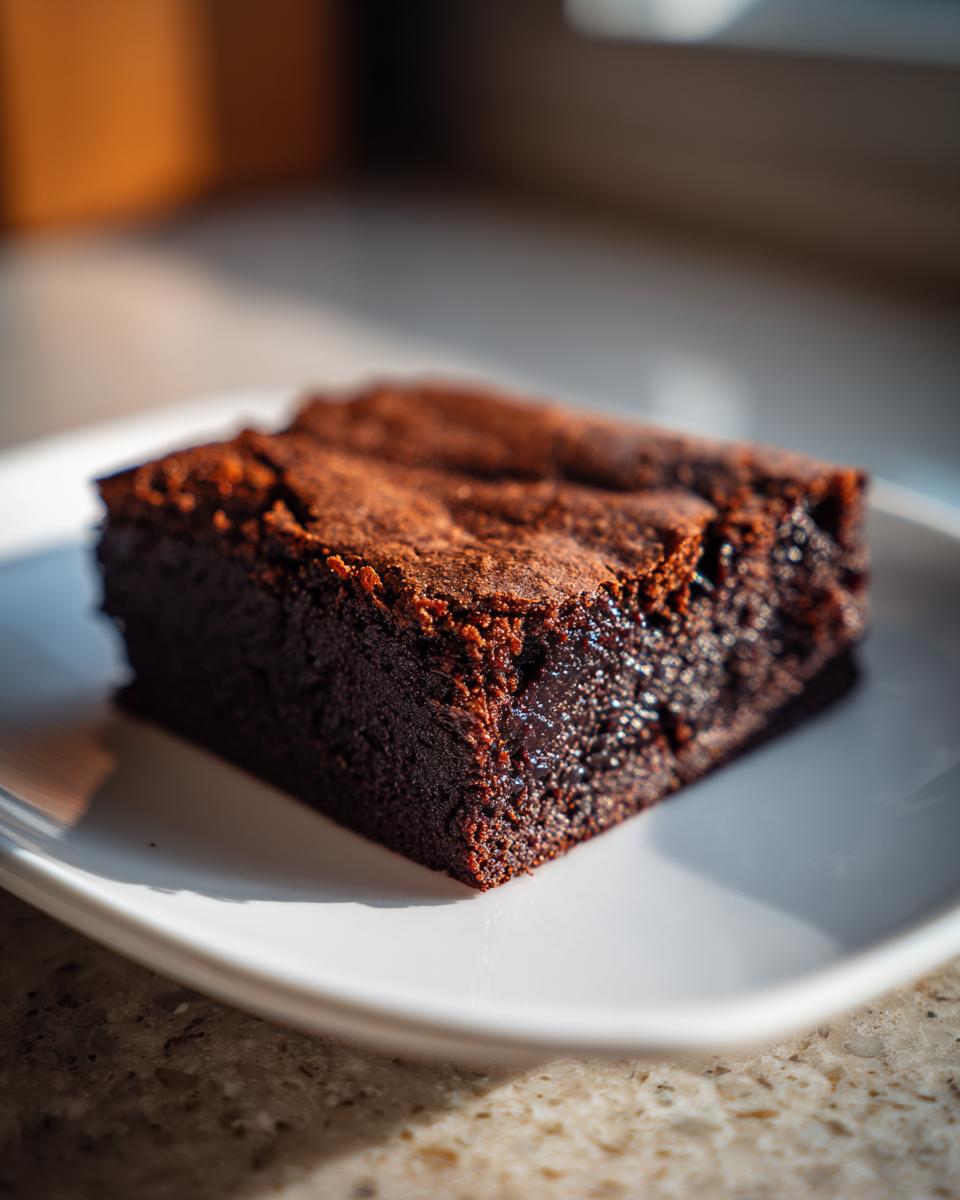 A close-up of a single, rich, fudgy square of date brownies resting on a white plate, highlighted by natural light.