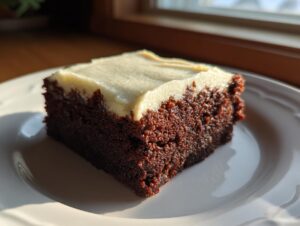 A close-up of a rich, dark slice of gingerbread brownies topped with thick, pale cream cheese frosting, sitting on a white plate.