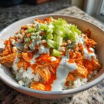 A close-up view of a delicious buffalo chicken bowl featuring shredded chicken coated in orange sauce, drizzled with ranch, topped with blue cheese crumbles and diced celery over rice.