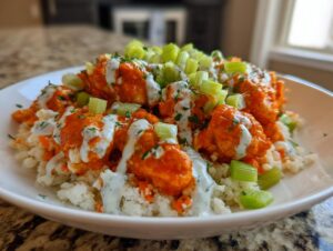 Close-up of a white bowl filled with a buffalo chicken bowl, featuring orange buffalo-sauced chicken over cauliflower rice, drizzled with ranch, and topped with celery.