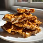 A stack of golden brown, glossy pieces of homemade peanut brittle recipe loaded with whole peanuts, sitting on a white plate.