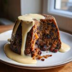 A rich, dark slice of plum pudding covered in flowing vanilla custard sauce on a white plate.