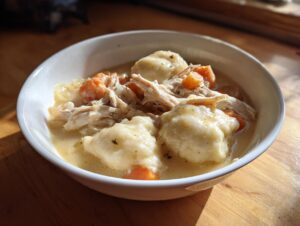 A close-up of a white bowl filled with creamy chicken and dumplings, shredded chicken, and visible carrots.