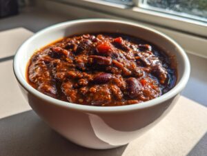 A close-up of a hearty bowl of rich, dark red black bean chili, bathed in sunlight near a window.