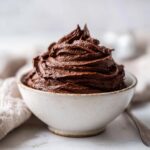 A close-up of rich, dark chocolate frosting beautifully piped into a swirl inside a small, speckled white bowl.