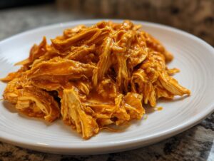 Close-up of shredded buffalo chicken coated in bright orange sauce piled on a white plate.