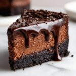 A rich slice of chocolate cheesecake topped with glossy chocolate ganache drizzle and cookie crumbs.